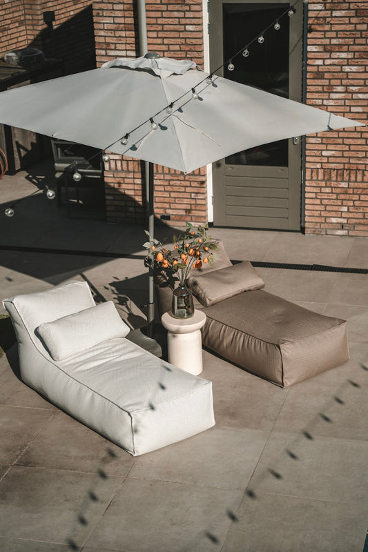 Outdoor patio with beige lounge chairs, a small table, and a large umbrella on a tiled floor.